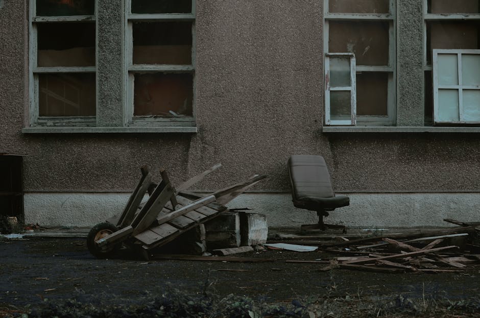 An outdoor scene showing a damaged, abandoned house with three broken, boarded-up windows in a weathered, dark brown exterior wall. In front of the house, on the pavement, there is a tipped-over wooden pallet and a small, white brick base with debris scattered around. To the right, an office-type swivel chair with a black seat and backrest is positioned on the pavement. Nearby, there are loose wooden planks and broken pieces of wood, indicating disrepair or disposal, possibly related to house clearance or waste removal. The lighting is natural, suggesting daytime, and the environment appears neglected, with signs of structural damage or demolition. The scene aligns with topics of house removals or clearance services, similar to those provided by Man with Van Harold Wood, especially in preparation for home relocation or waste disposal, supporting the context of removal solutions for avoiding bulky waste fines in Harold Wood.