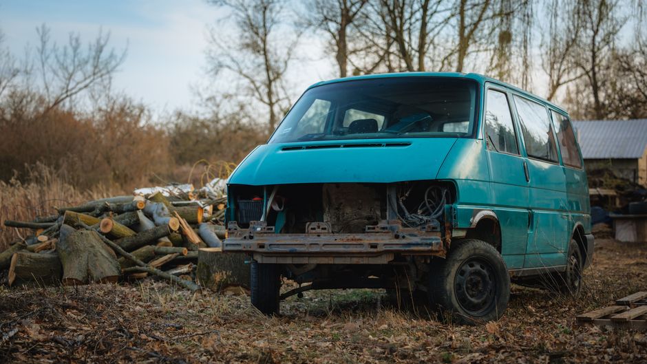 A weathered, turquoise van with no front bumper and visible rust on its exposed metal frame is parked outdoors on uneven ground covered with dry grass, leaves, and debris. The van's windshield and sid