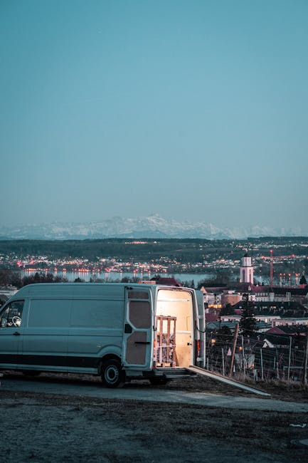 A weathered, turquoise van with no front bumper and visible rust on its exposed metal frame is parked outdoors on uneven ground covered with dry grass, leaves, and debris. The van's windshield and sid