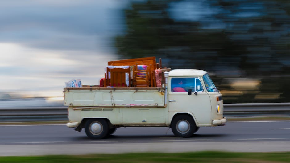 A vintage cream-colored van with an open cargo area is shown moving along a roadway during daylight hours, with a blurred background indicating motion. The van's cargo space is filled with various household items, including wooden chairs, cardboard boxes, plastic-wrapped packages, and cushions, arranged to maximize storage. The vehicle is positioned in the centre of the image, and the scene suggests the process of a home relocation. The background features a guardrail and indistinct trees or buildings, emphasizing speed and transport. The image illustrates the type of vehicle used by Man with Van Harold Wood for furniture transport and packing and moving services involving house removals from Gubbins Lane to Brentwood Road in Harold Wood, highlighting the practical aspects of loading and moving furniture during a property transfer.