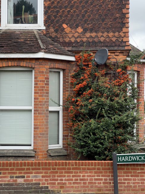 Image of a brick residential building with two large windows featuring white blinds and a bay window on the ground floor. To the right, a dense shrub with red and green foliage partially covers the wall, growing near a satellite dish attached to the exterior. Blue sky is visible above the roof, which has brown tiles. In the lower right corner, a green street sign reading 'HARDWICK' is mounted on a black post. The scene is well-lit, capturing elements typical of a home relocation environment, with a focus on the exterior features of the property, suitable for illustrating house removals or moving logistics in Harold Wood. Man with Van Harold Wood’s services involve careful moving and packing of household items, often requiring access to such properties for loading furniture and belongings onto moving vehicles, which are visible outside or nearby in the broader context of a quiet residential street.