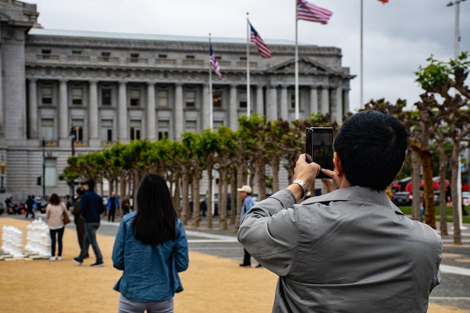 A man with short black hair, wearing a grey jacket, is holding a smartphone in both hands and taking a photo of a large grey government building with classical architectural features, tall columns, and flags flying on flagpoles outside. In the foreground, a woman with long black hair, dressed in a blue jacket, stands on a paved area with landscaped trees and shrubs nearby. Several other pedestrians are walking across the open space, some facing the building and others in various directions. The scene appears to be during daytime under overcast weather, with a mix of paved and landscaped surfaces surrounding the building, possibly indicating a city or town square. The image reflects a scene of public observation, possibly related to city planning, historic architecture, or local government, aligning with themes of relocation, transport, and move logistics. As part of Man with Van Harold Wood's service for house removals, this image highlights the urban environment and logistical considerations involved in home relocation and furniture transport.