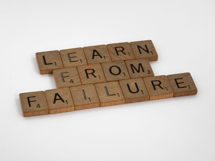 A close-up image shows three rows of wooden Scrabble tiles arranged on a white surface, each row spelling out words related to moving: 'LEARN', 'FROM', and 'FAILURE'. The tiles are light brown with black letterings and small numbers indicating their point value. The background is plain and bright, emphasizing the tiles. This visual could symbolize the challenges faced during a home relocation or furniture transport process. The image is clean and well-lit, highlighting the textual arrangement, and subtly relates to the concepts of learning from past experiences and overcoming obstacles in house removals, fitting with services provided by Man with Van Harold Wood for efficient packing and moving logistics.
