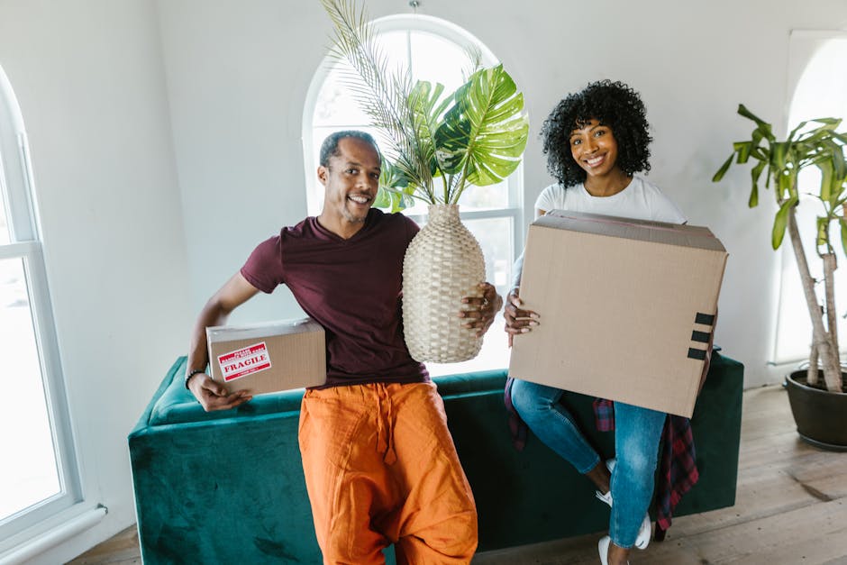 A man and woman are indoors in a bright, modern room with natural light coming through an arched window. The man, wearing a maroon t-shirt and orange trousers, is smiling and holding a small cardboard box marked fragile in his right hand. He is also holding a large, woven ceramic vase with a green leafy plant inside it, supported by his left hand. The woman, dressed in a white top and blue jeans with a plaid shirt tied around her waist, is seated on a dark green sofa and holding a large cardboard box, likely packed for a home relocation. The room features wooden flooring, a tall potted plant in a black pot on the right, and the atmosphere suggests preparation for packing or moving as part of a furniture transport or packing and moving process. This scene exemplifies household packing activities during local house removals, as facilitated by [COMPANY_NAME].