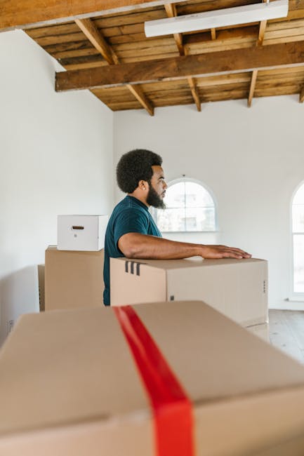 A man with curly black hair and a beard, wearing a dark blue T-shirt, is inside a spacious room with white walls and a wooden ceiling. He is standing behind large cardboard moving boxes, some stacked on top of each other, with the top box having a red packing strap. The room has two tall, arched windows allowing natural light to illuminate the space, with a view of outdoor buildings visible through the windows. The boxes are positioned near a doorway, and the man appears to be preparing for a home relocation, possibly involved in packing or moving furniture as part of a house removal. This scene exemplifies furniture transport and packing during a local move, with visible cardboard boxes and a clean, organized environment typical of moving and clearance services provided by Man with Van Harold Wood.