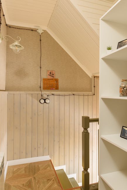 Inside a residential property showing a staircase with carpeted steps and a wooden handrail, leading downward to a lower level. The upper wall features textured beige wallpaper, with a wooden framed artwork and a small wooden wall mount near the ceiling. A vintage-style ceiling light with a glass shade is visible, hanging from a cord. To the right, a white shelving unit displays framed photographs and decorative items. The corner of the lower staircase area shows the edge of a wooden staircase with a patterned wood floor partially visible. This scene is captured during a home relocation or packing process, as part of furniture transport or moving logistics handled by Man with Van Harold Wood, evident from the natural lighting and arranged interior environment aimed at easing the moving process during house removals in Harold Wood.