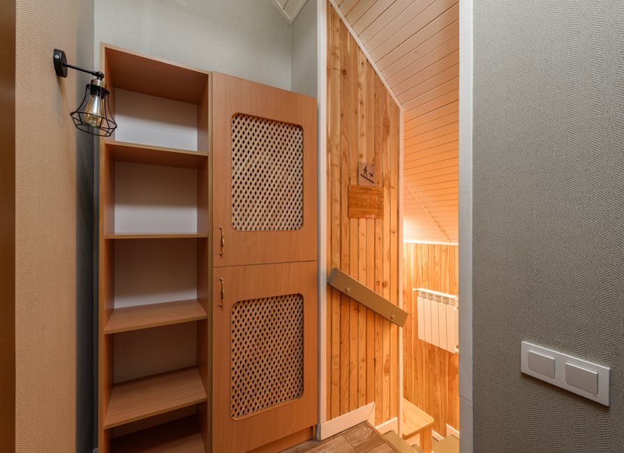 The image shows an interior view of a residential property featuring a wooden storage cabinet with two lattice-patterned doors and open shelves on the left side, positioned against a light-colored wall. To the left of the cabinet, a decorative black wall-mounted light fixture with an exposed bulb is visible. In the background, a staircase with wooden paneling and a beige handrail leads downwards, with a cardboard box resting on the top step, indicating ongoing packing or furniture transport as part of a home relocation. The surrounding environment includes a textured wall on the right with a double light switch, and the ceiling has wood paneling, contributing to the warm and functional interior ambiance. The scene appears to be part of a moving process managed by Man with Van Harold Wood, demonstrating the preparation and logistical aspects of furniture transport through narrow stairs in Harold Wood flats.