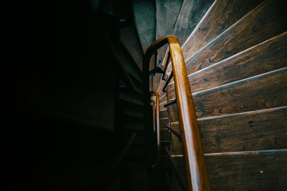 Inside a residential property showing a staircase with carpeted steps and a wooden handrail, leading downward to a lower level. The upper wall features textured beige wallpaper, with a wooden framed artwork and a small wooden wall mount near the ceiling. A vintage-style ceiling light with a glass shade is visible, hanging from a cord. To the right, a white shelving unit displays framed photographs and decorative items. The corner of the lower staircase area shows the edge of a wooden staircase with a patterned wood floor partially visible. This scene is captured during a home relocation or packing process, as part of furniture transport or moving logistics handled by Man with Van Harold Wood, evident from the natural lighting and arranged interior environment aimed at easing the moving process during house removals in Harold Wood.