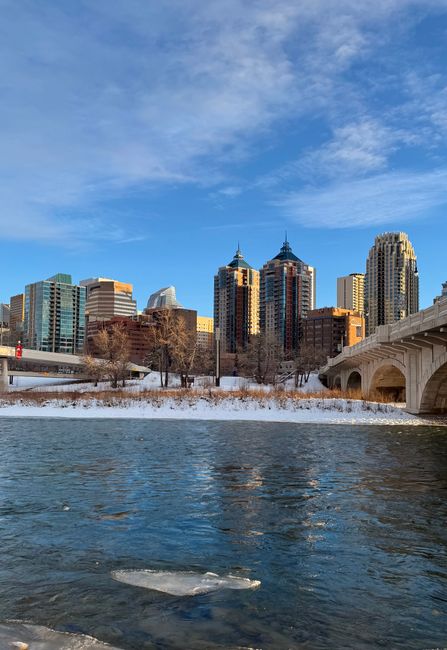A photograph taken during daytime shows the riverbank in Harold Wood with snow-covered ground and leafless trees lining the water's edge. In the background, there is a large city skyline with numerous tall buildings made of glass and concrete, some with pointed rooftops, under a partly cloudy blue sky. On the right side of the image, part of a stone bridge with arches extends over the water. Near the riverbank, visible objects include cardboard boxes, wrapped furniture covered with plastic sheeting or moving blankets, and a hand truck or trolley being used to transport these items during a house removal process. Slightly inside the property or near the pavement, a person is seen carrying a box, and another is loading items onto a van. The scene suggests active loading or packing as part of a relocation carried out by Man with Van Harold Wood, supporting the logistics involved in home relocations or furniture transport services.