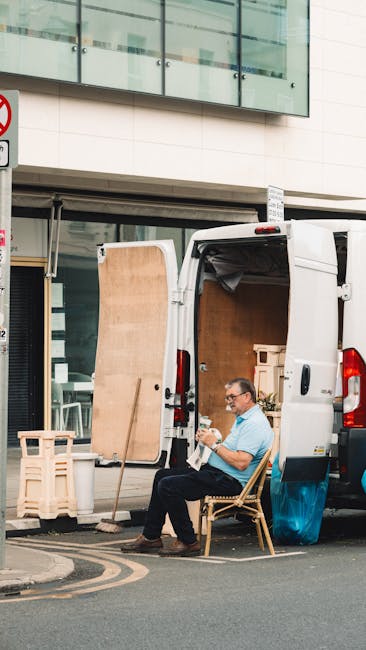 A man wearing glasses and a light blue shirt sits on a wooden chair outside a white delivery van parked adjacent to a modern building with large glass windows and a white facade. The rear doors of the